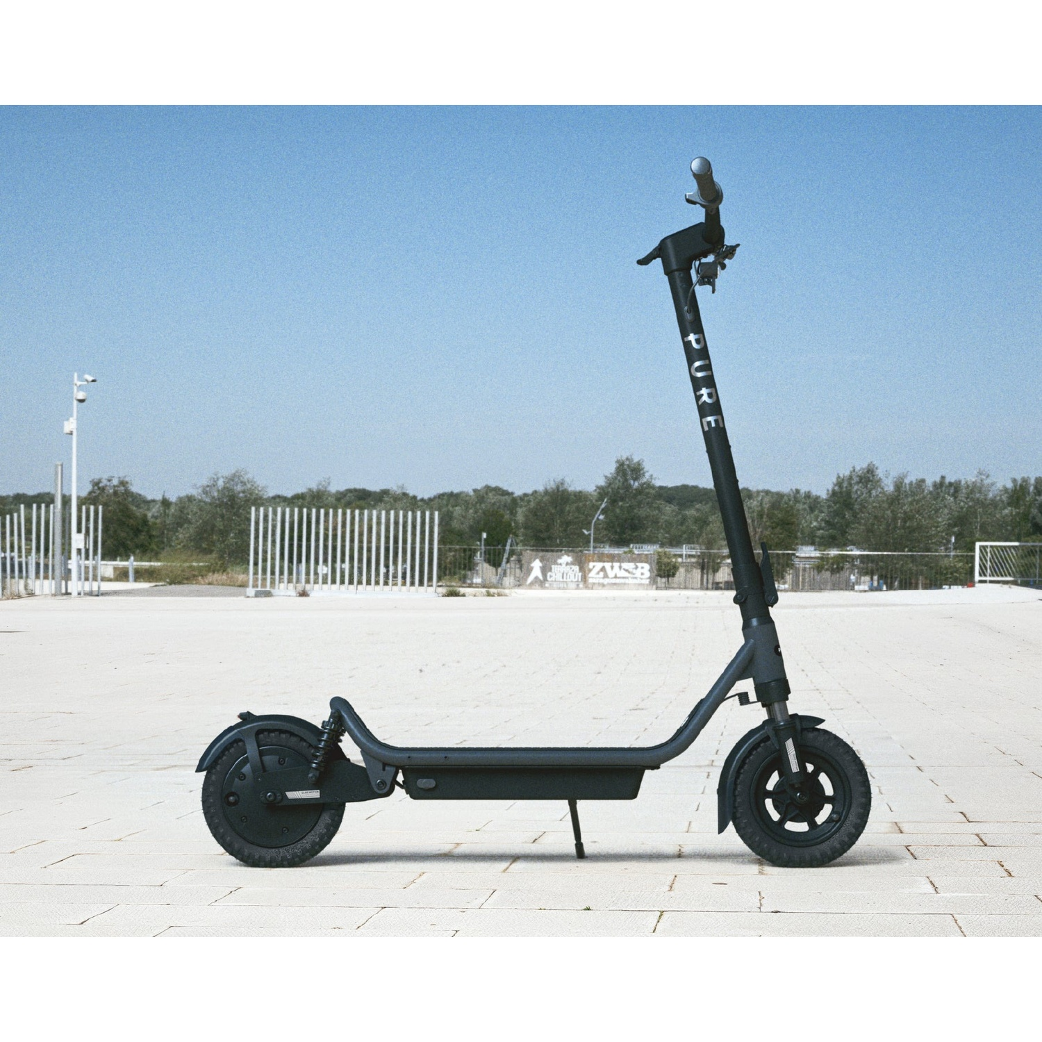 A black electric scooter parked on a paved square with trees and a clear blue sky.