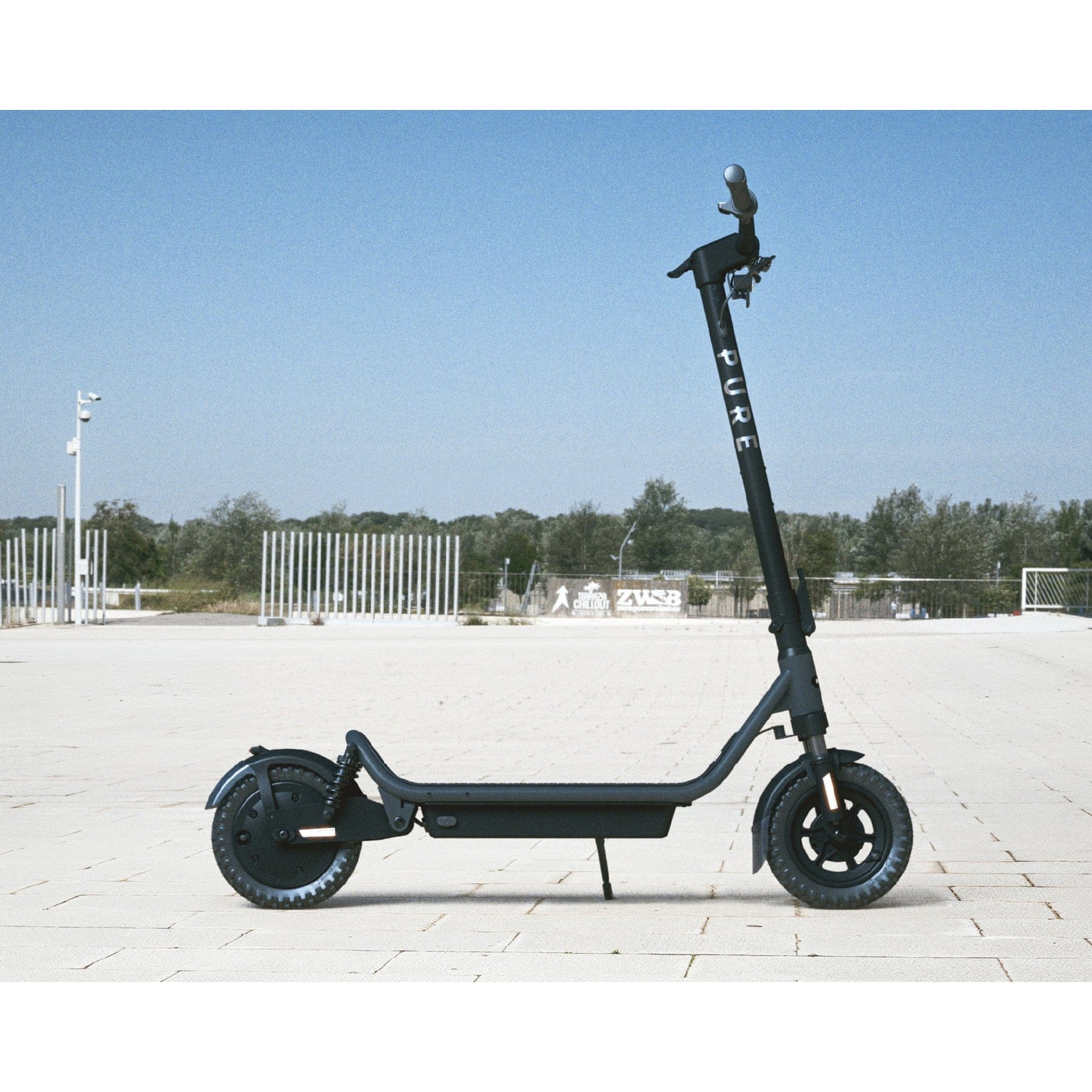 A black electric scooter parked outdoors on a paved square against trees and a clear blue sky.