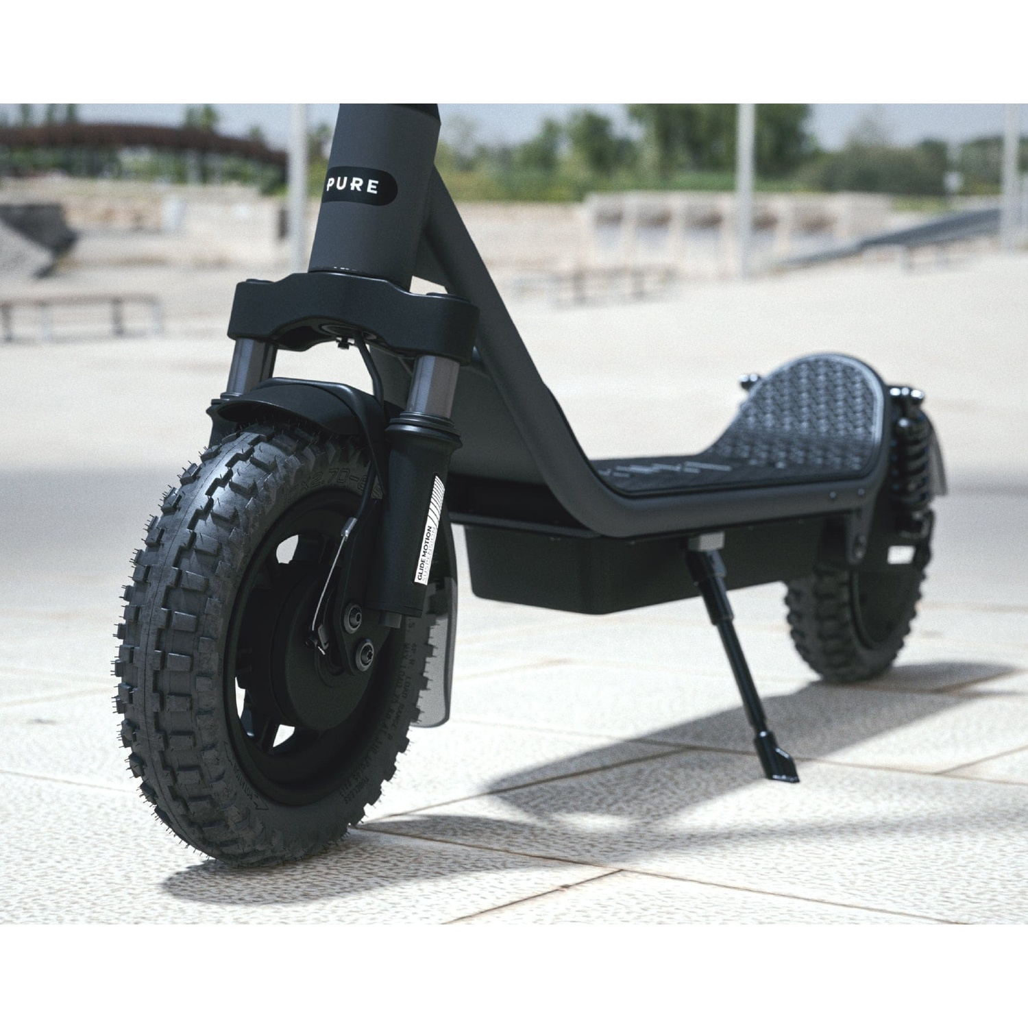 A black Pure electric scooter with large off-road tires parked on a light stone plaza with trees behind.