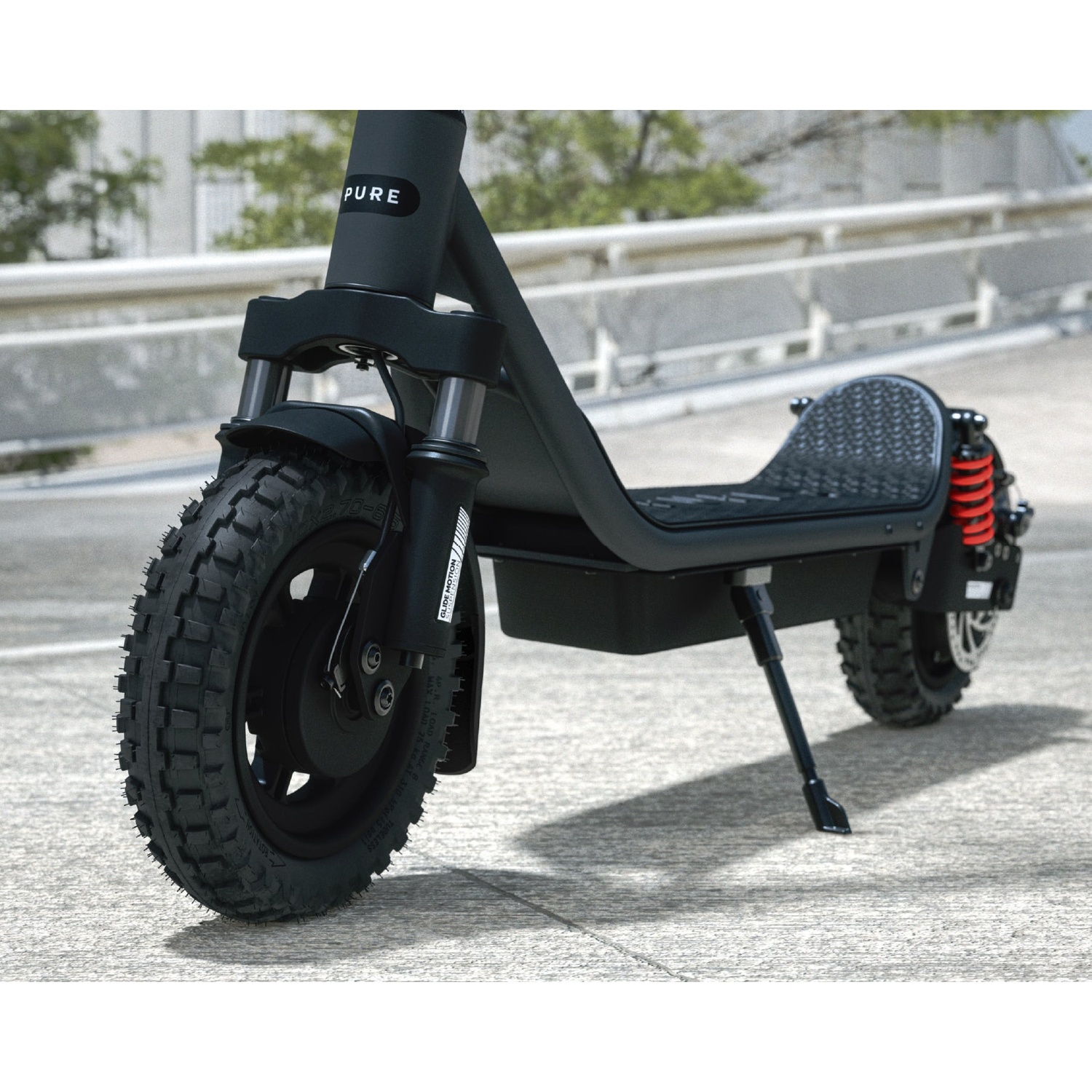 A black electric scooter with knobby tires and red suspension stands on a bright ground in front of a white wall.