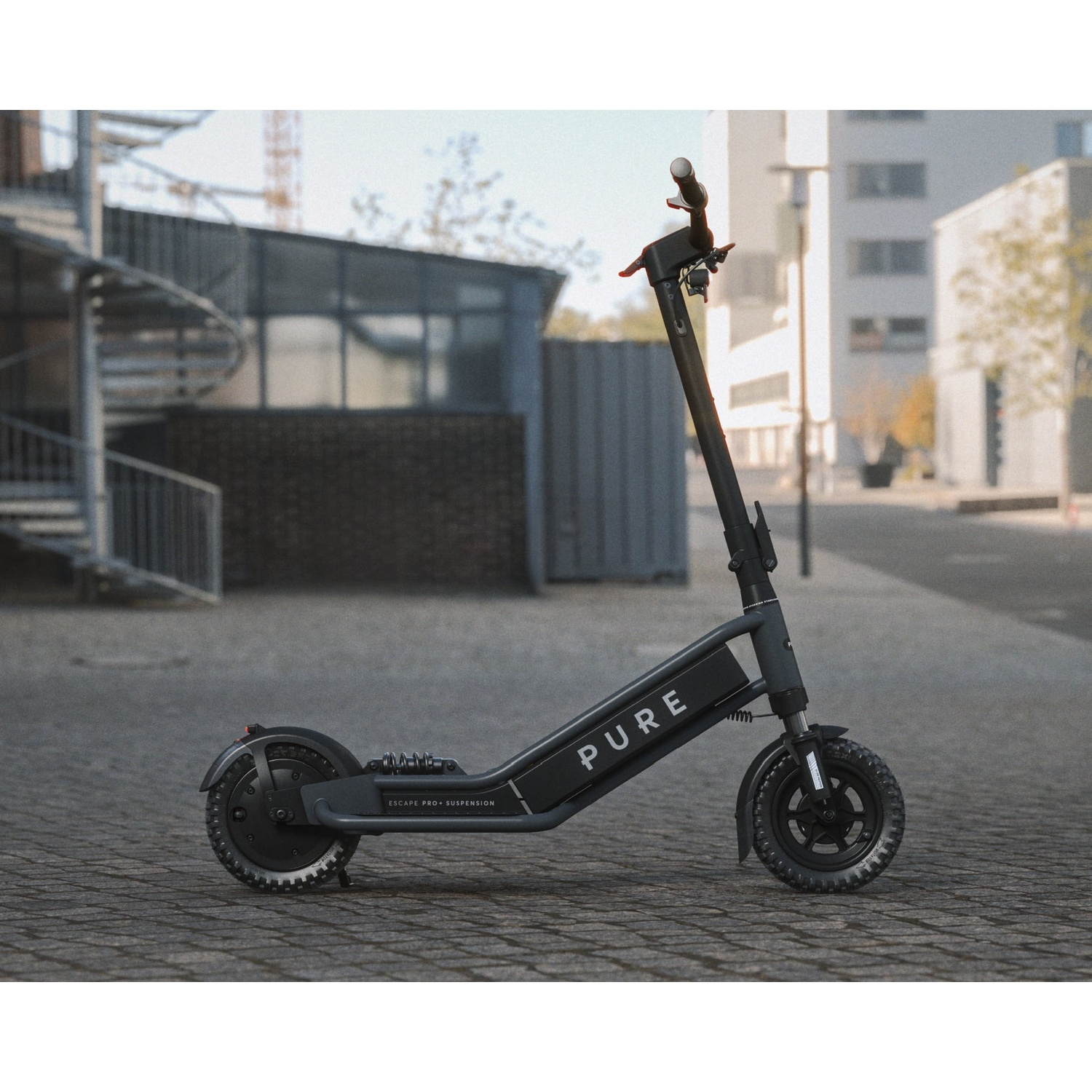 A gray Pure electric scooter stands on a cobblestone street in a city, with modern buildings in the background.