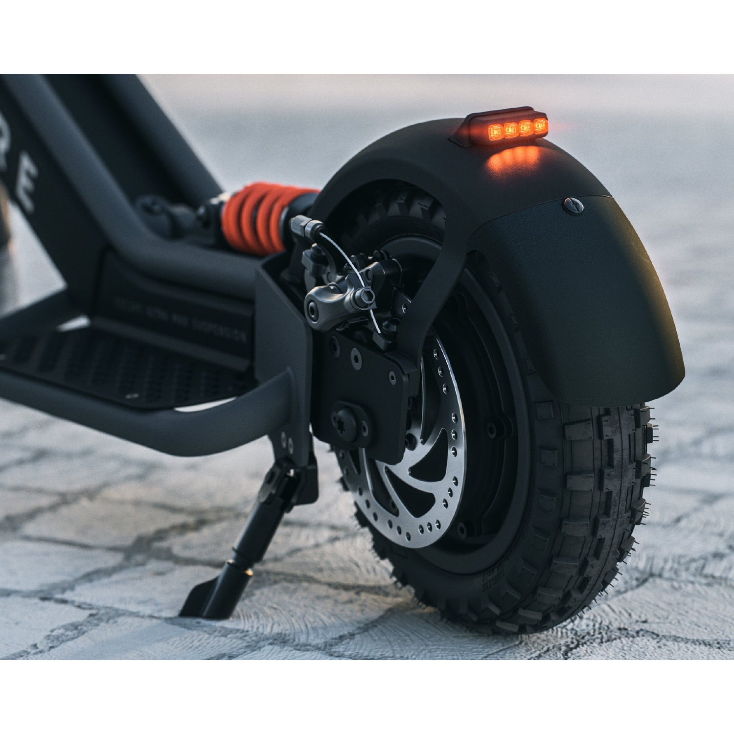 Close-up of a black electric scooter's rear wheel, disc brake, red suspension spring, and lit tail light on pavement.