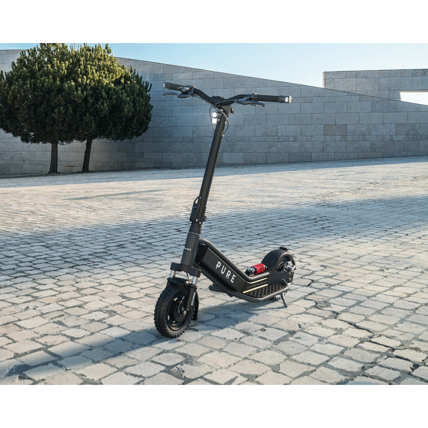A black electric scooter stands on a stone plaza in front of a modern concrete wall and a small tree.