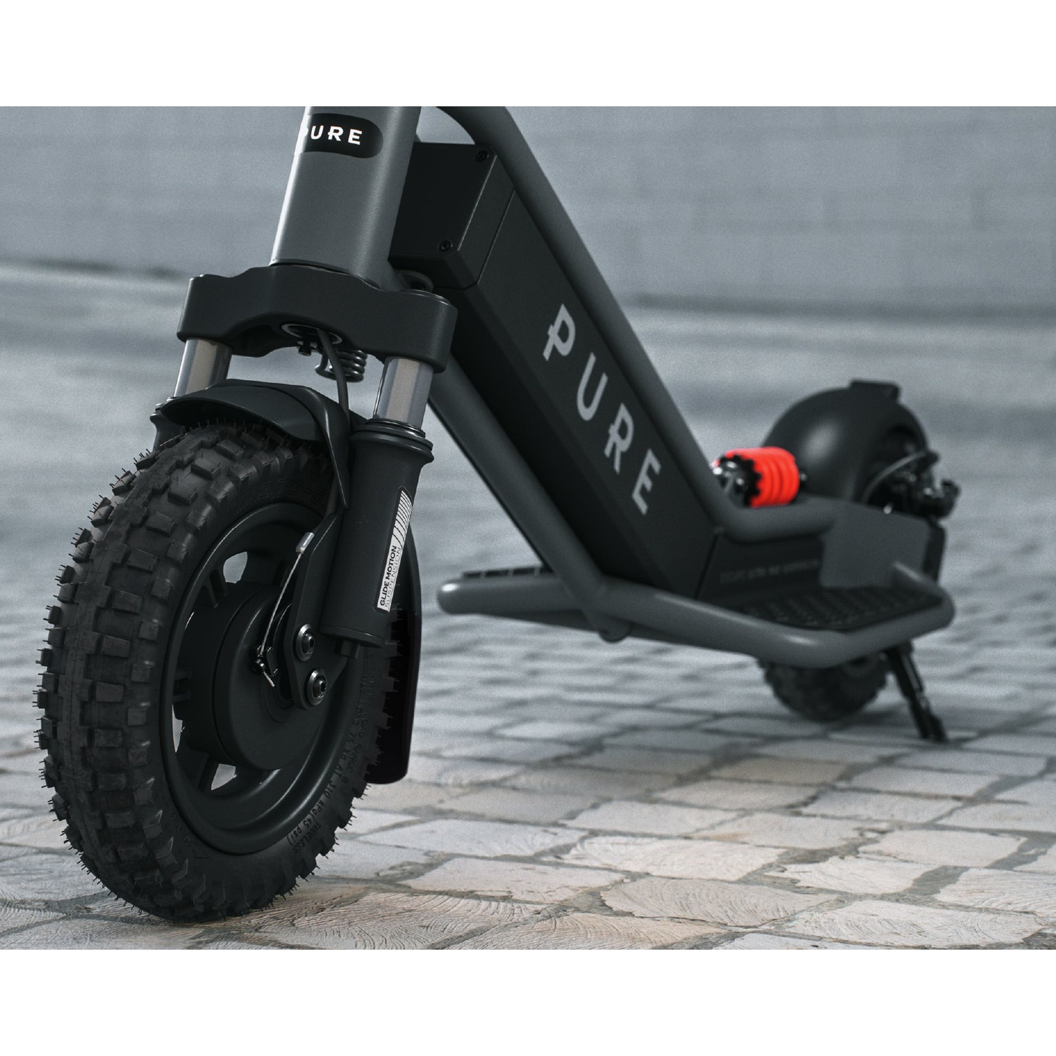 A dark gray Pure electric scooter with knobby tires parked on a cobblestone surface.