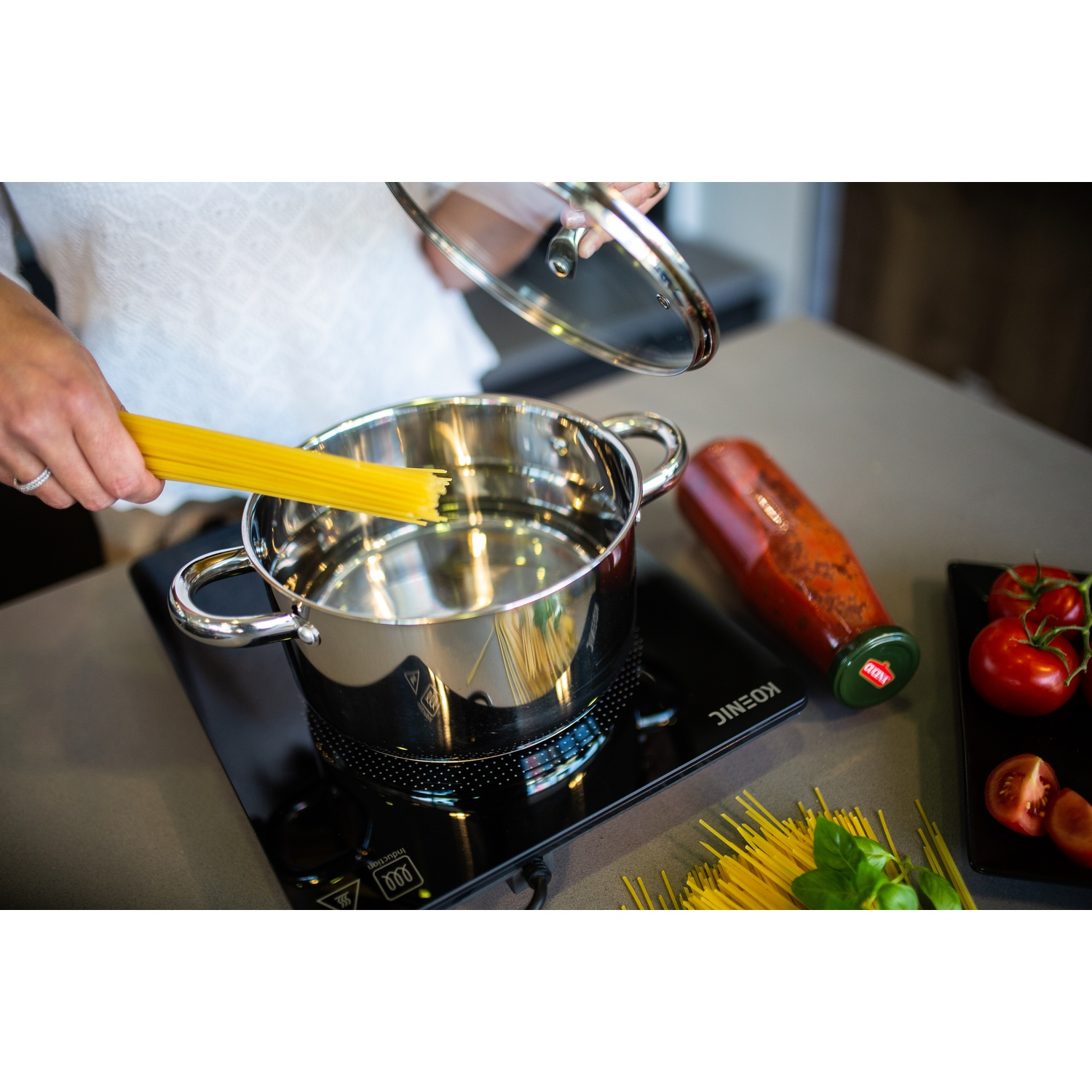 A person placing spaghetti into a pot on an induction cooktop next to tomatoes and a sauce bottle.