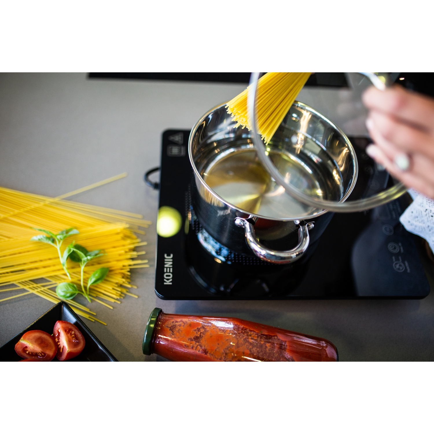 A pot on an induction cooktop as spaghetti is added, with tomatoes and basil nearby.