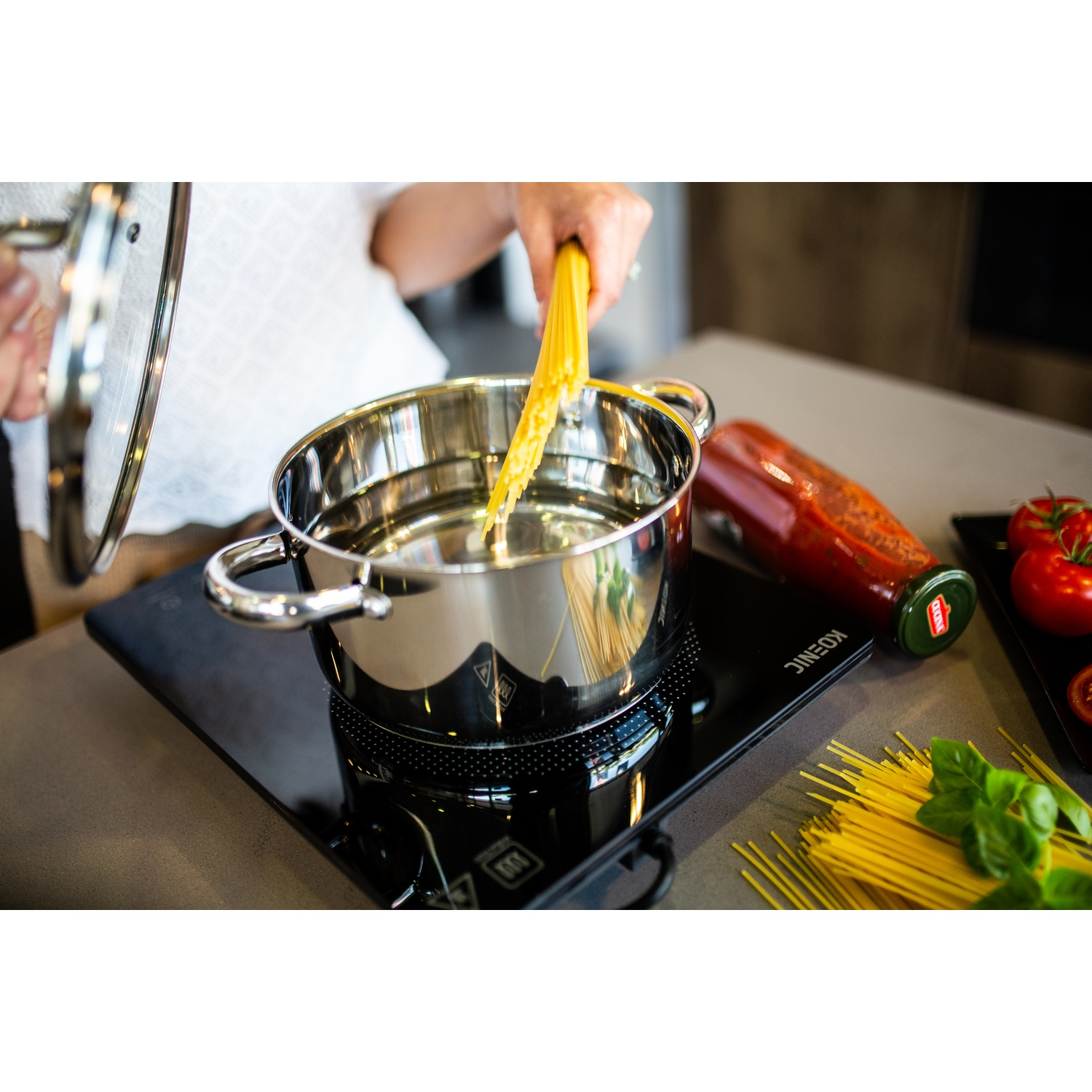 A person placing raw spaghetti into a boiling pot of water on a black induction cooktop in a kitchen.