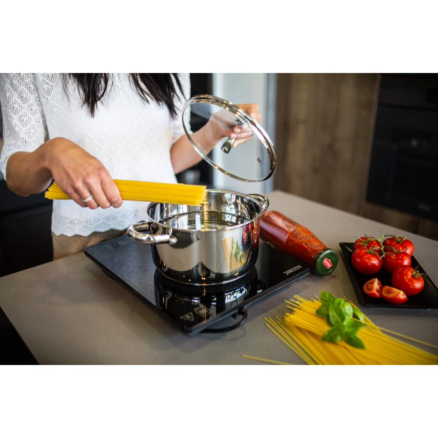 Woman holding lid and pasta over a pot of water on a stove, with tomatoes nearby.