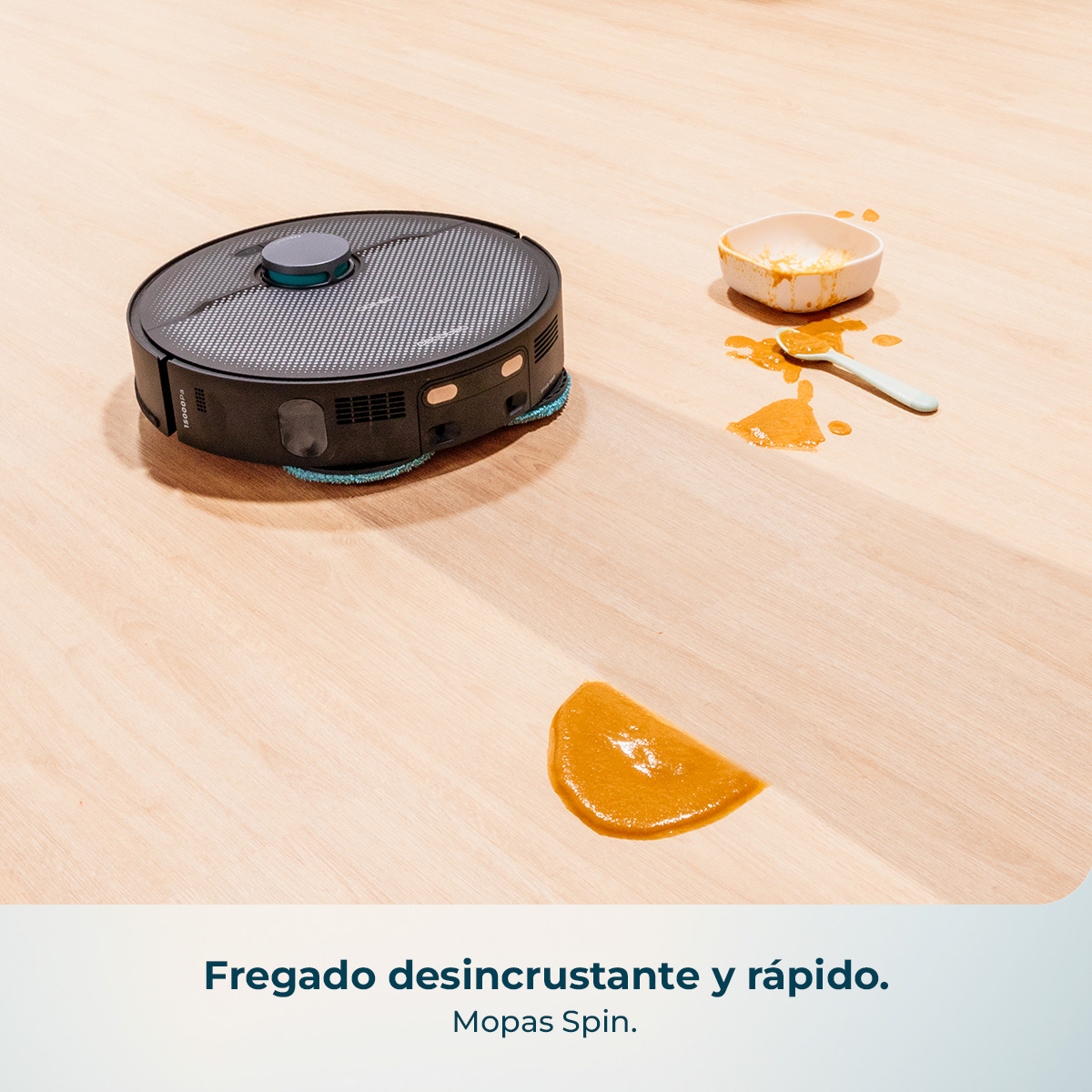 Un robot aspirador negro se encuentra en un suelo de madera cerca de una comida derramada.