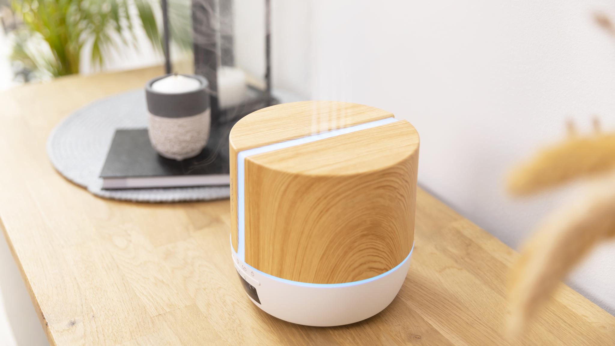 Un difusor de aroma cilíndrico de madera con luz azul brillante está sobre una mesa de madera, emitiendo vapor.