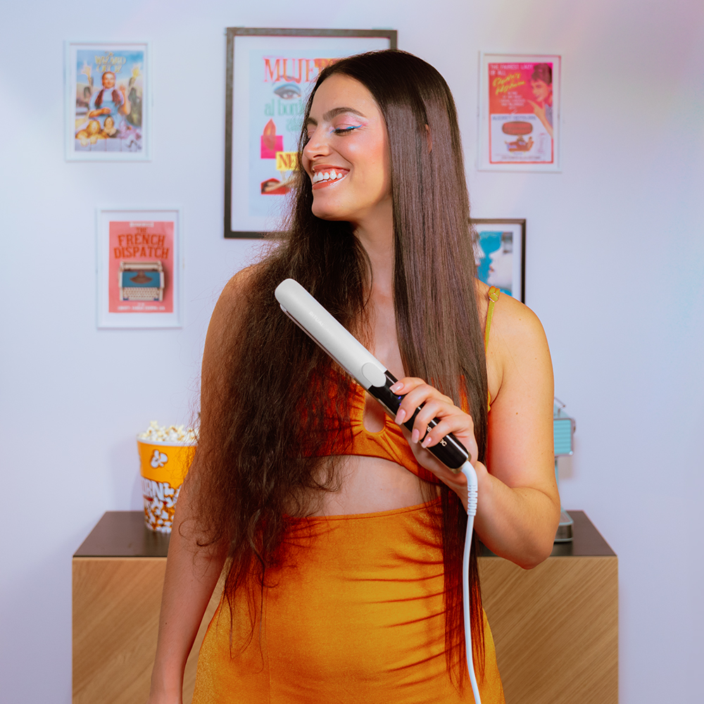Una mujer sonriente con vestido naranja sostiene una plancha de pelo ante carteles de cine.