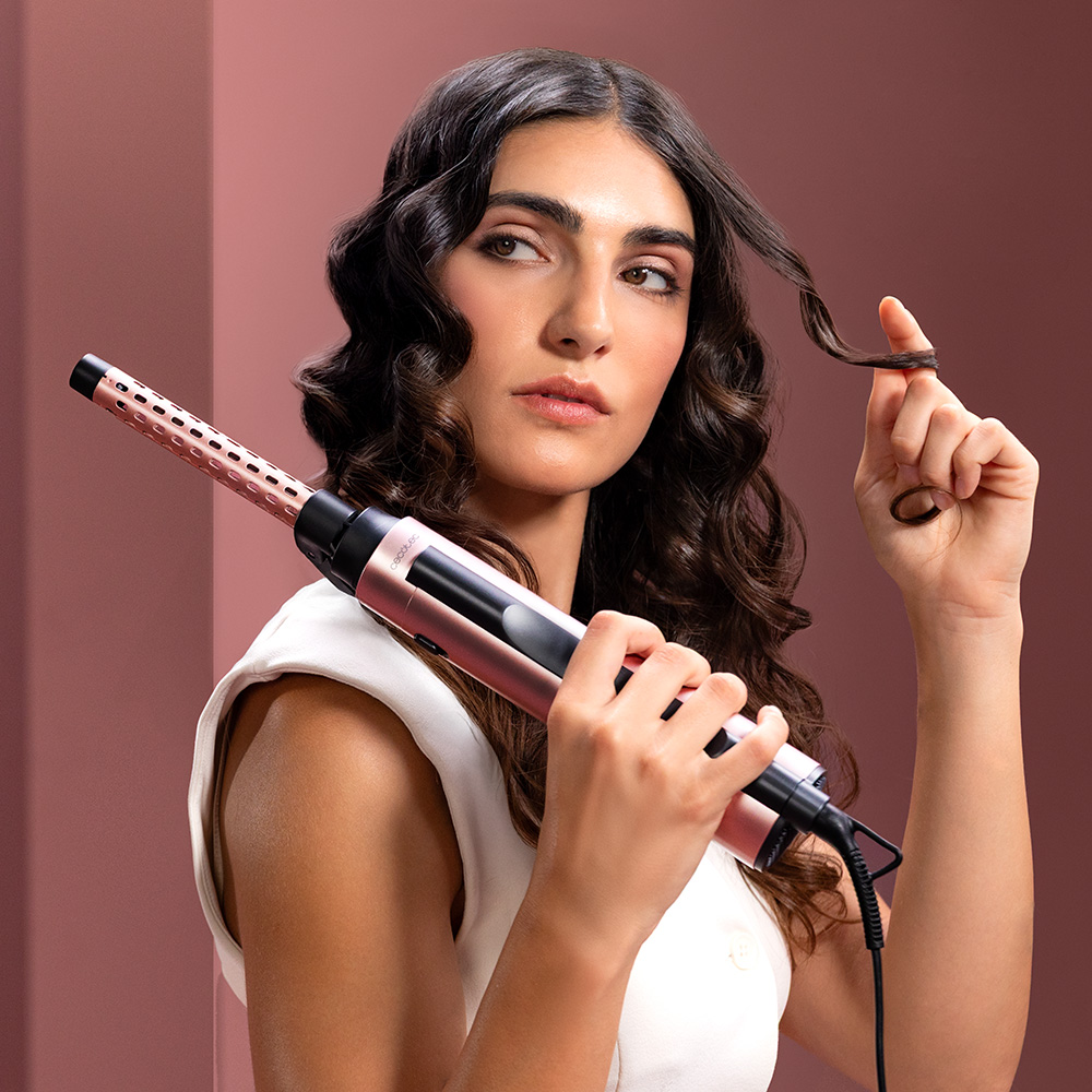 Una mujer con cabello oscuro y rizado sostiene un rizador de oro rosa ante una pared morada.