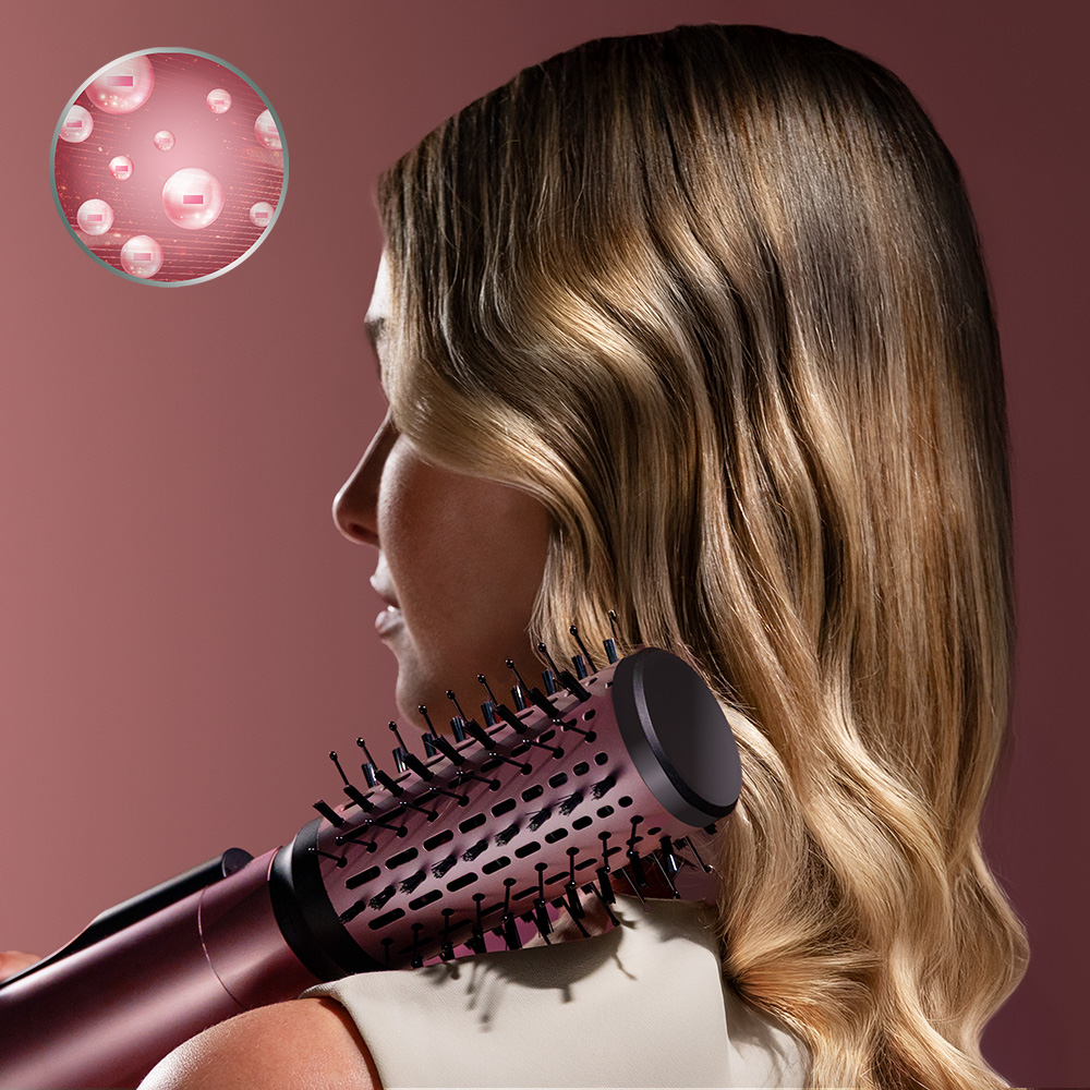 Una mujer rubia sostiene un cepillo estilizador rosa junto al hombro, con un icono circular que muestra iones negativos.