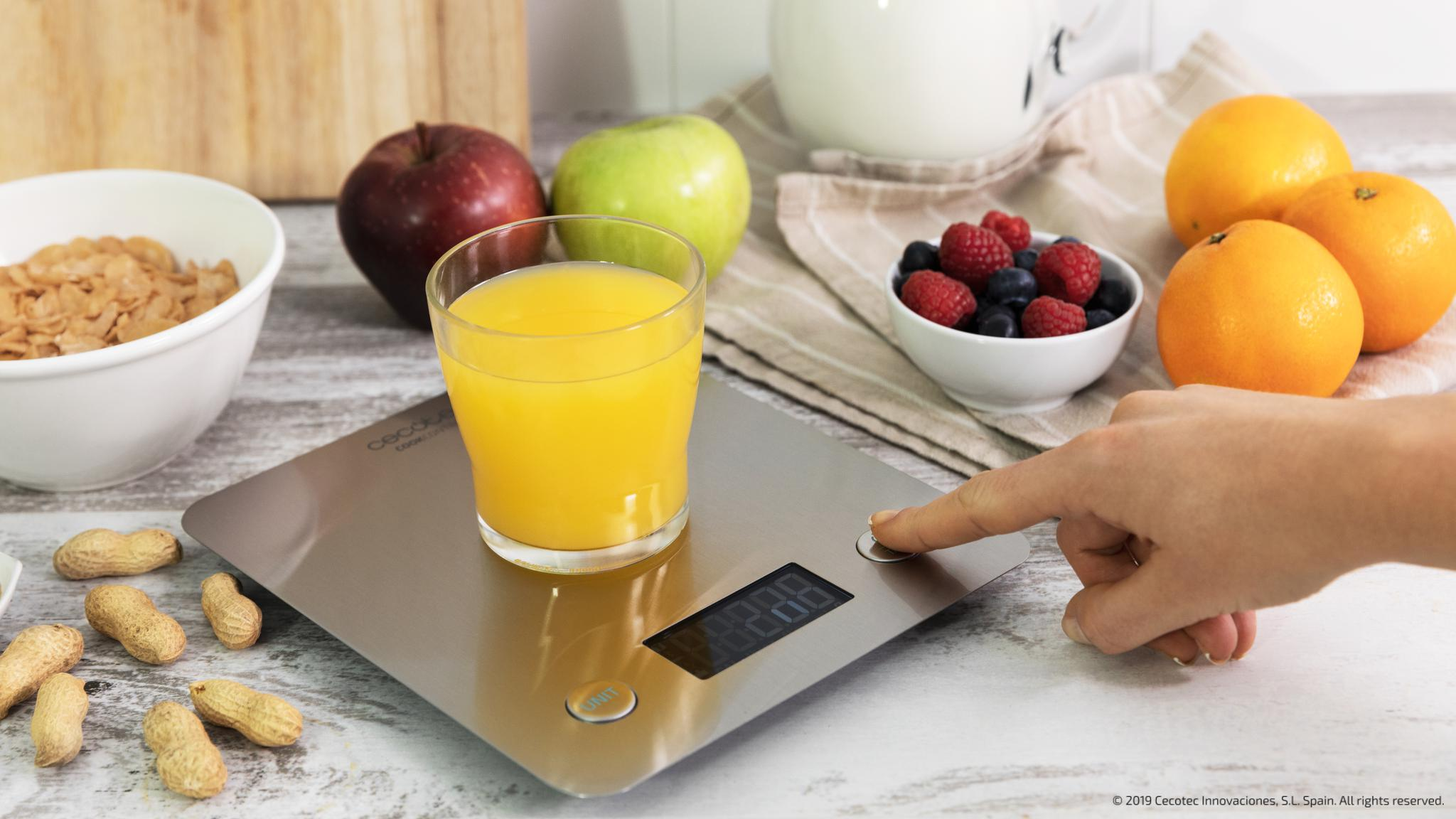 Una mano presiona el botón de encendido de una báscula de cocina plateada con zumo de naranja, rodeada de fruta.