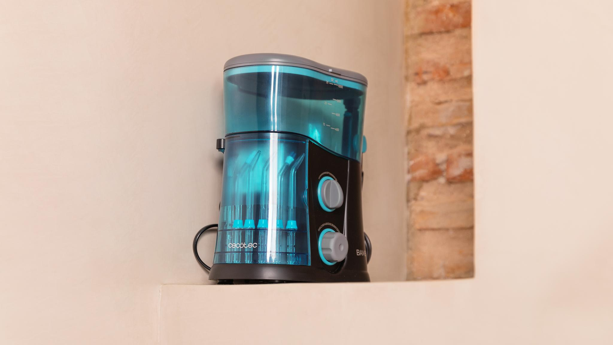 Un irrigador bucal azul y negro con accesorios se encuentra en un estante contra una pared beige.