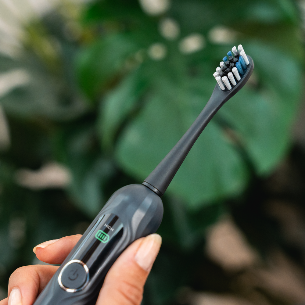 Una mano sostiene un cepillo de dientes eléctrico oscuro frente a hojas verdes.