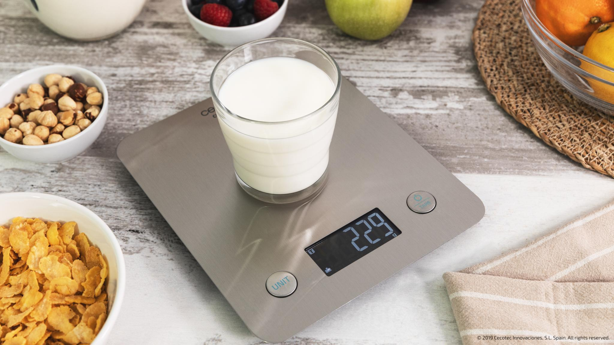 Un vaso de leche en una báscula de cocina digital, rodeado de tazones con cereales, frutos secos y bayas en una mesa.