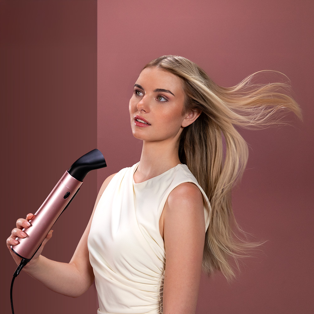 Una mujer rubia con vestido blanco sostiene un secador rosa ante fondo malva.