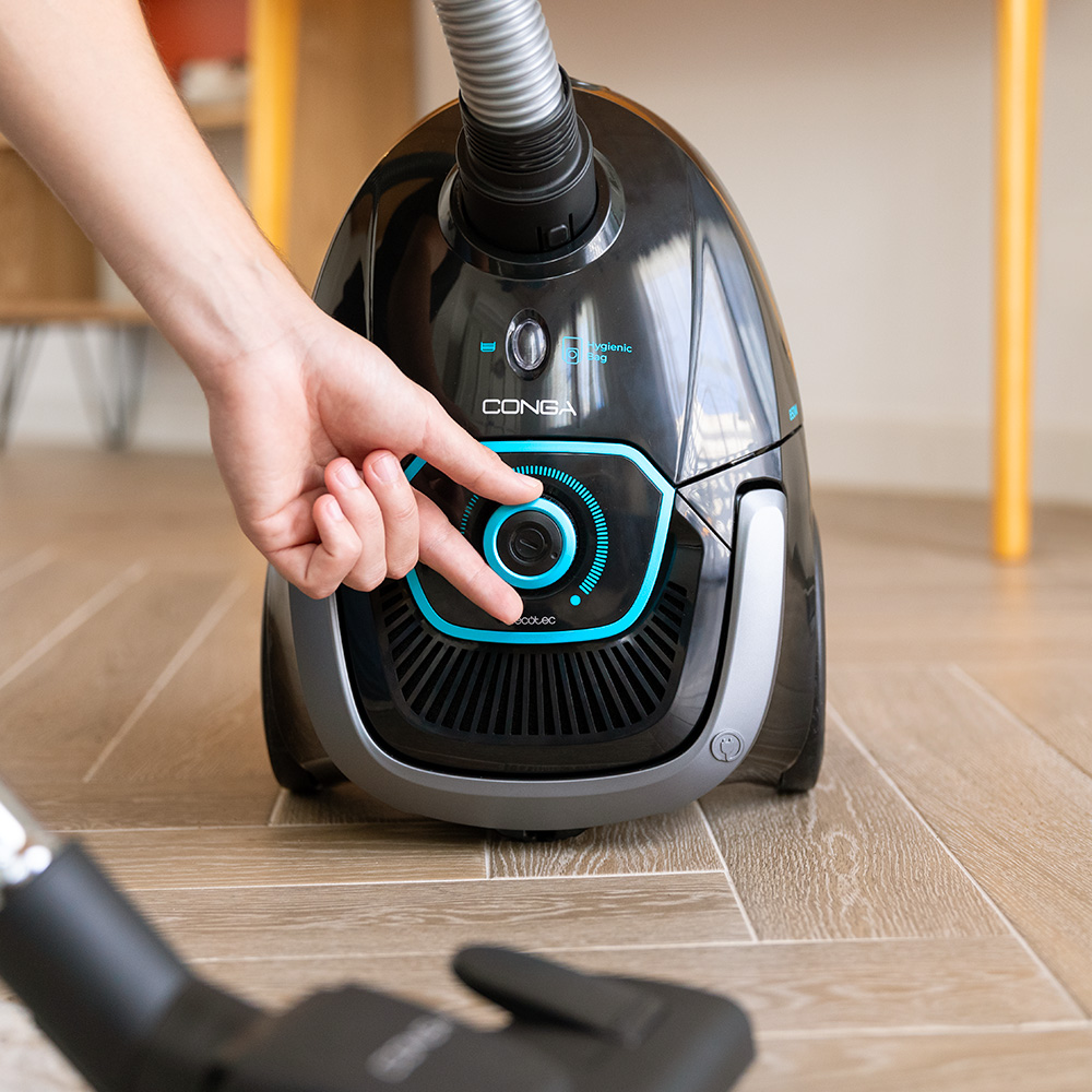 Una mano gira el dial azul de una aspiradora negra en un suelo de madera.