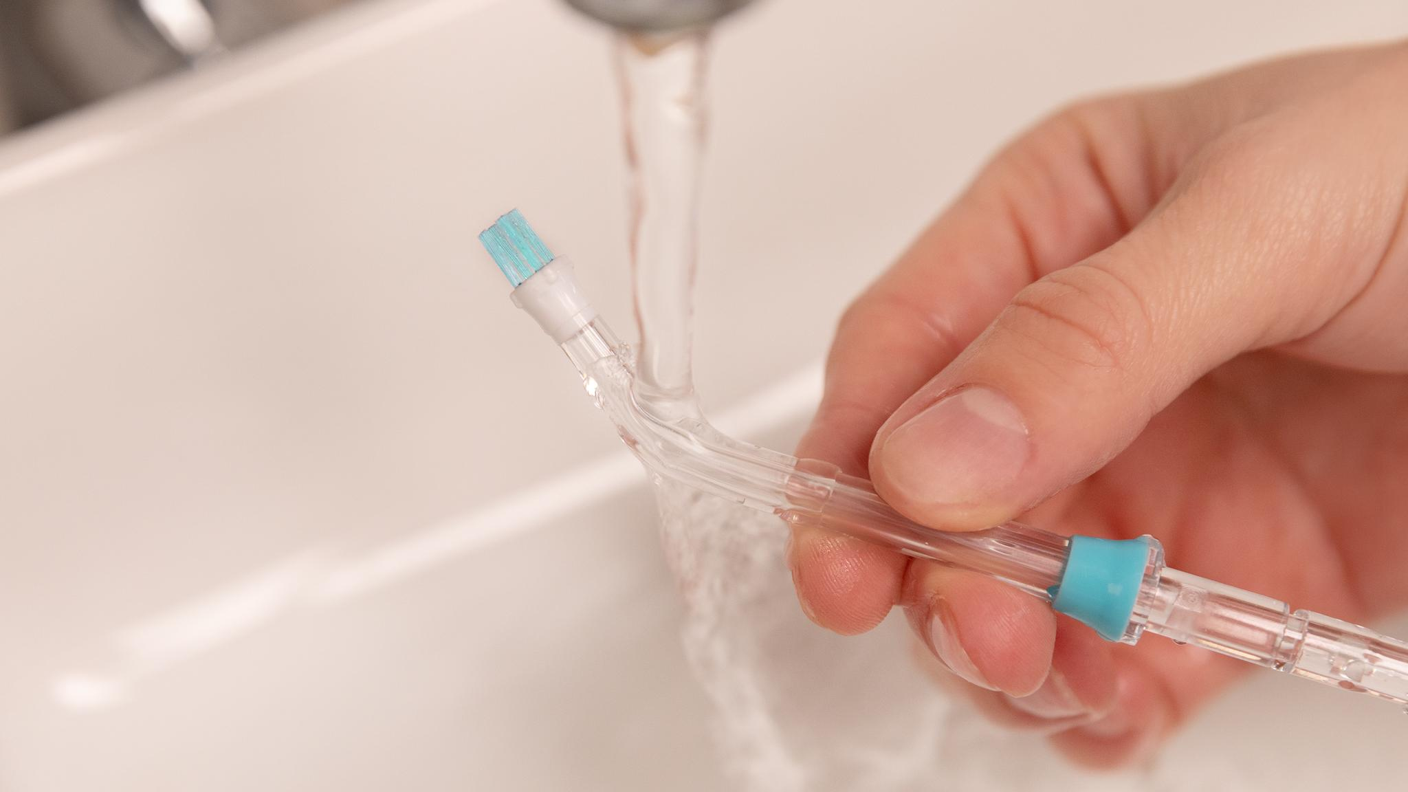 Una mano sostiene un tubo transparente con punta de cepillo azul bajo un grifo abierto.