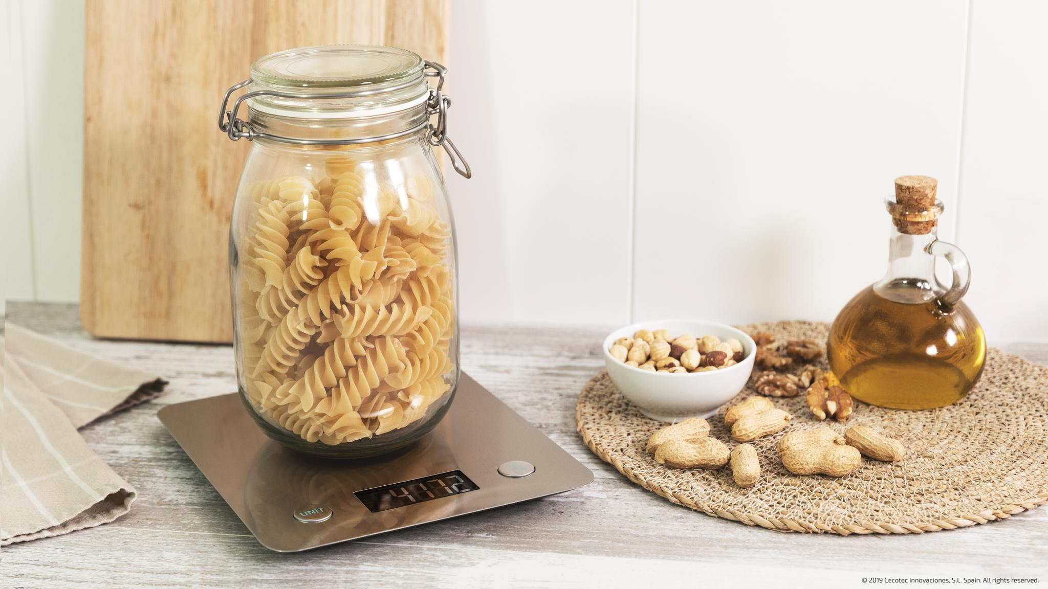 Un frasco de pasta fusilli en una báscula de cocina digital, con frutos secos y aceite cerca.
