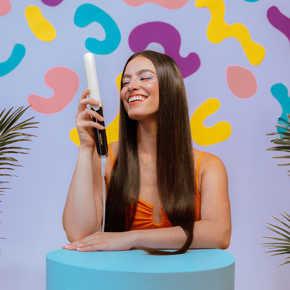 Una mujer sonriente con largo pelo castaño sostiene una plancha de pelo frente a un fondo colorido.