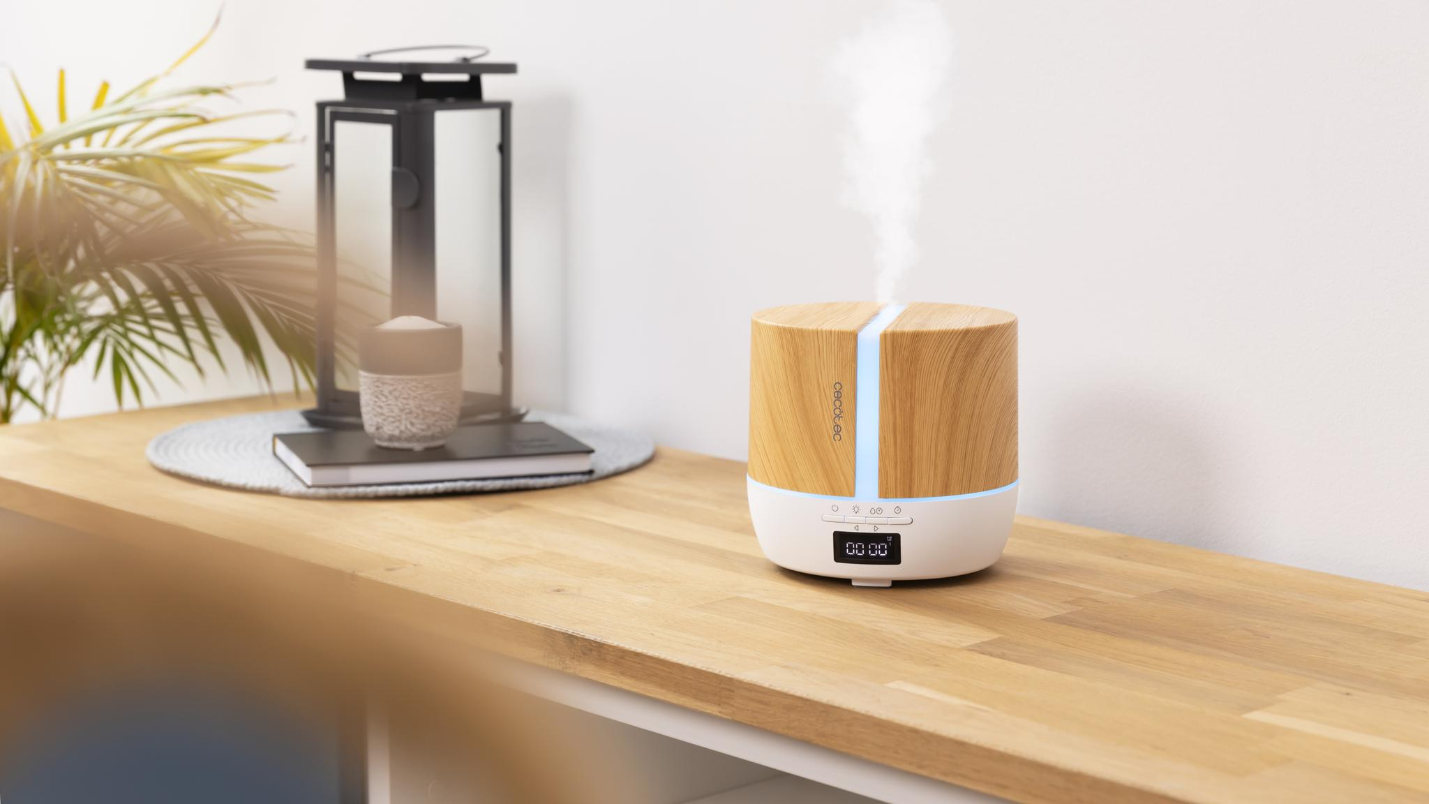 Un difusor de aroma de madera en una mesa emite vapor, con un farol y plantas de fondo.