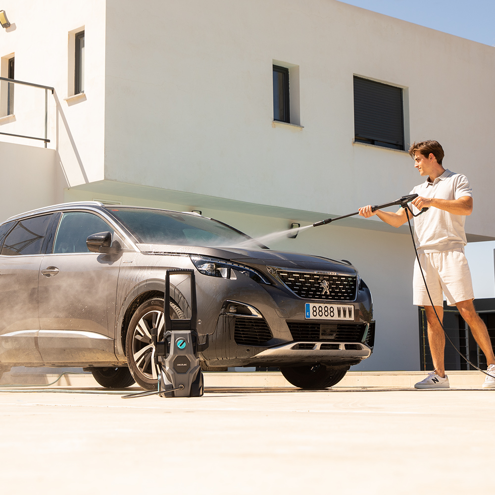 Un hombre lava un coche gris bajo el sol con una hidrolimpiadora frente a una casa blanca moderna.