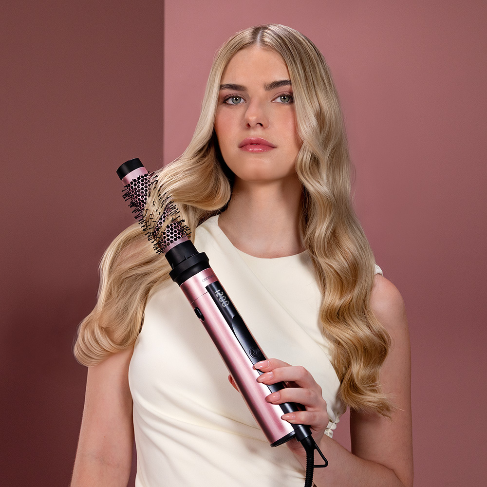 Una mujer rubia con top blanco sostiene un cepillo moldeador rosa y negro sobre fondo rosa.