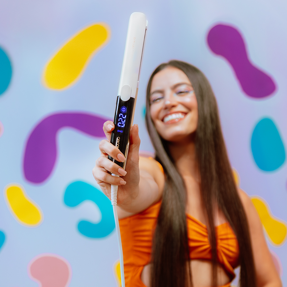 Una mujer sonriente sostiene una plancha de pelo blanca hacia la cámara frente a un fondo colorido y estampado.