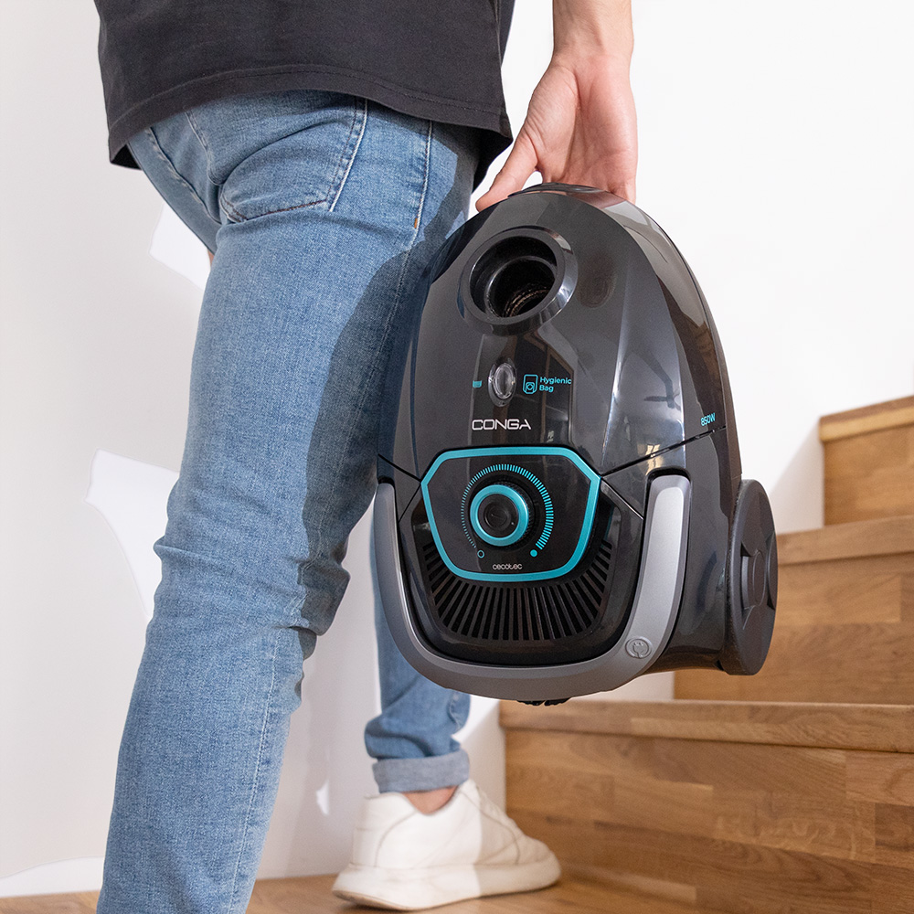 Una persona en jeans sostiene una aspiradora moderna gris y turquesa en escaleras de madera.