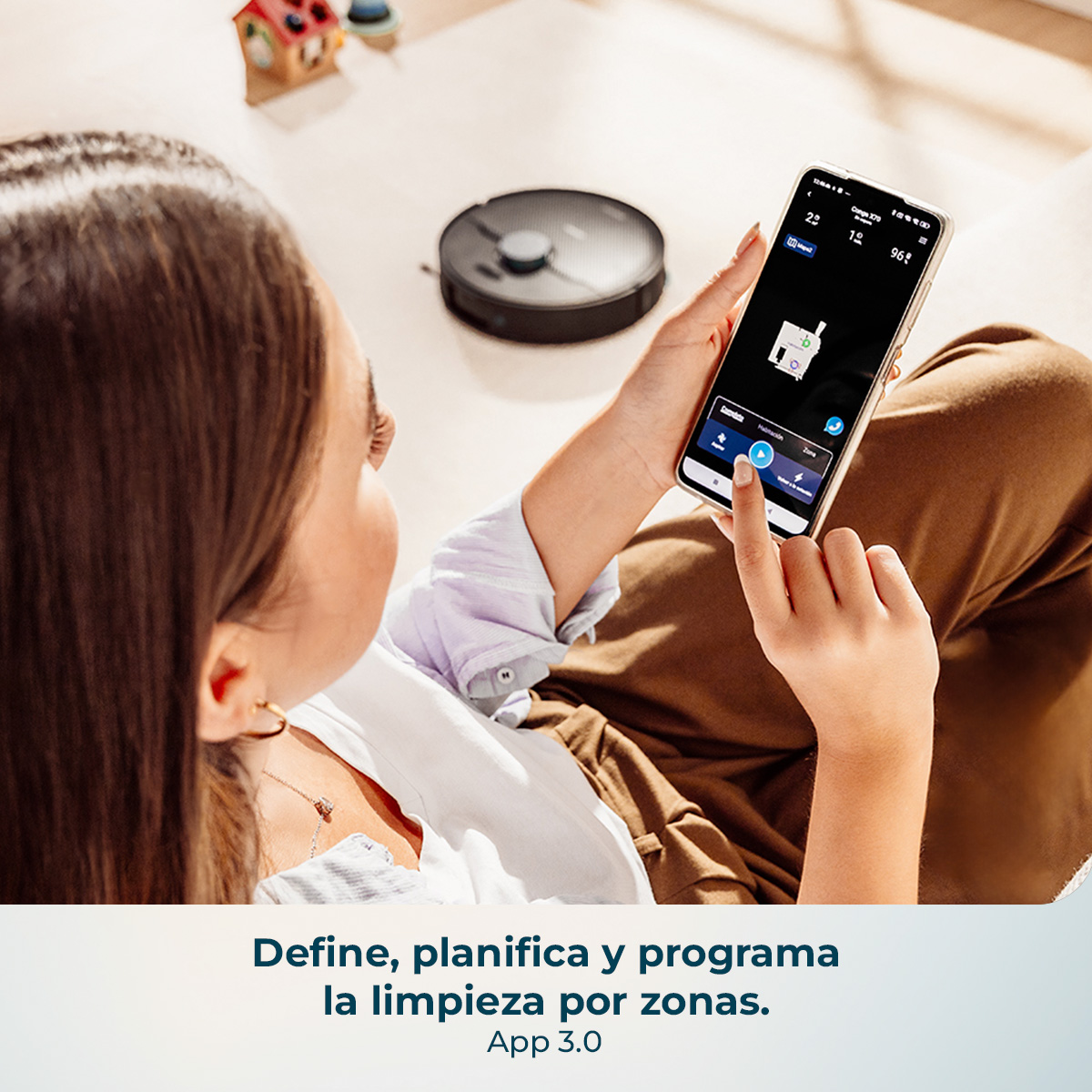 Una mujer usa una app en su smartphone para controlar un robot aspirador que está en el suelo cercano.