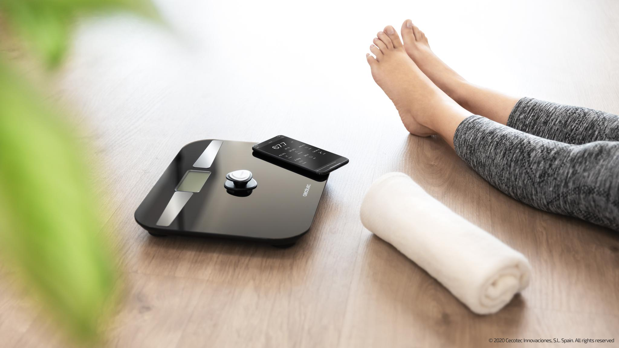 Una persona está sentada en el suelo junto a una báscula digital negra, un smartphone y una toalla blanca.