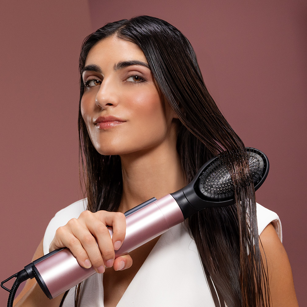 Una mujer con cabello largo y oscuro sostiene un cepillo moldeador de aire rosa dorado frente a un fondo rosa.