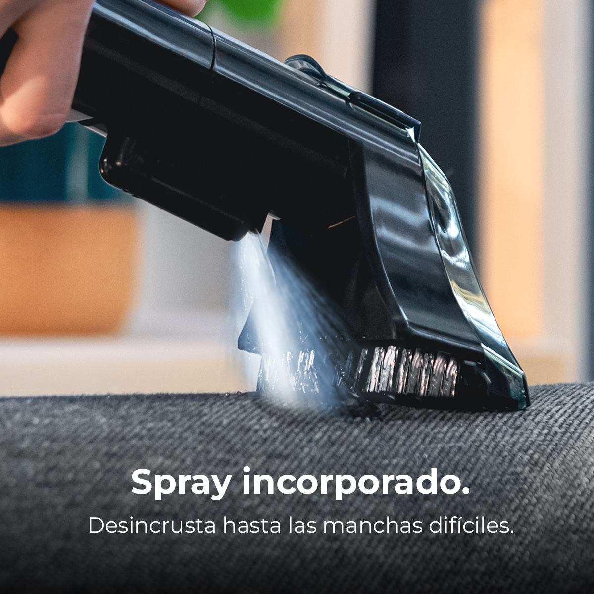 Mano sostiene limpiador negro pulverizando agua sobre un sofá gris.
