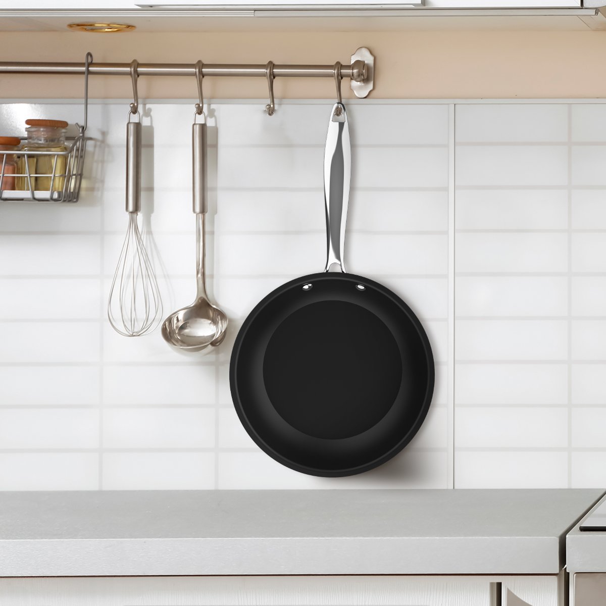 Una sartén negra y utensilios de cocina plateados cuelgan de una barra contra azulejos blancos.