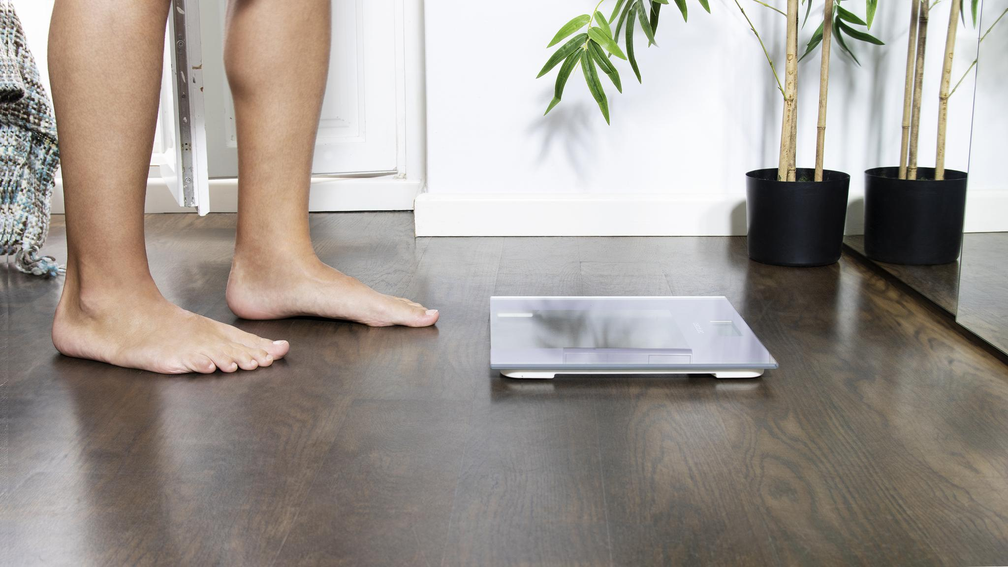 Persona descalza parada frente a una báscula de baño digital en un piso de madera, con una planta en maceta al fondo.
