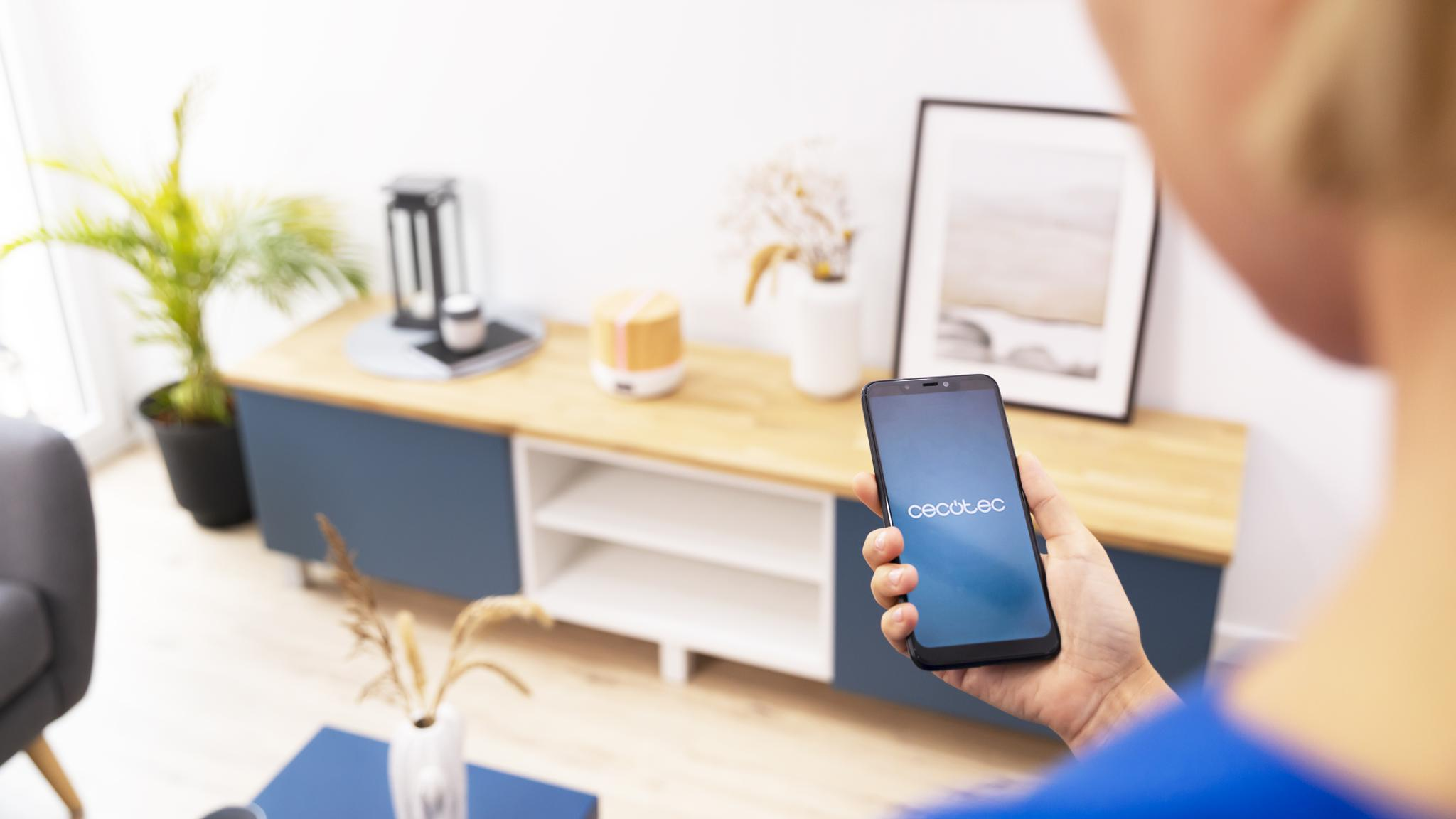 Persona sosteniendo un smartphone con el logotipo de Cecotec en un salón con mueble azul y blanco.