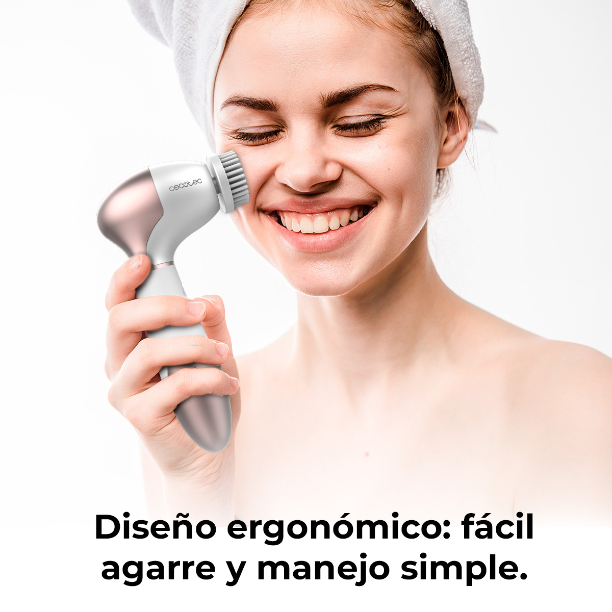 Una mujer sonriente con una toalla en el cabello sostiene un cepillo limpiador facial blanco y dorado en su mejilla.