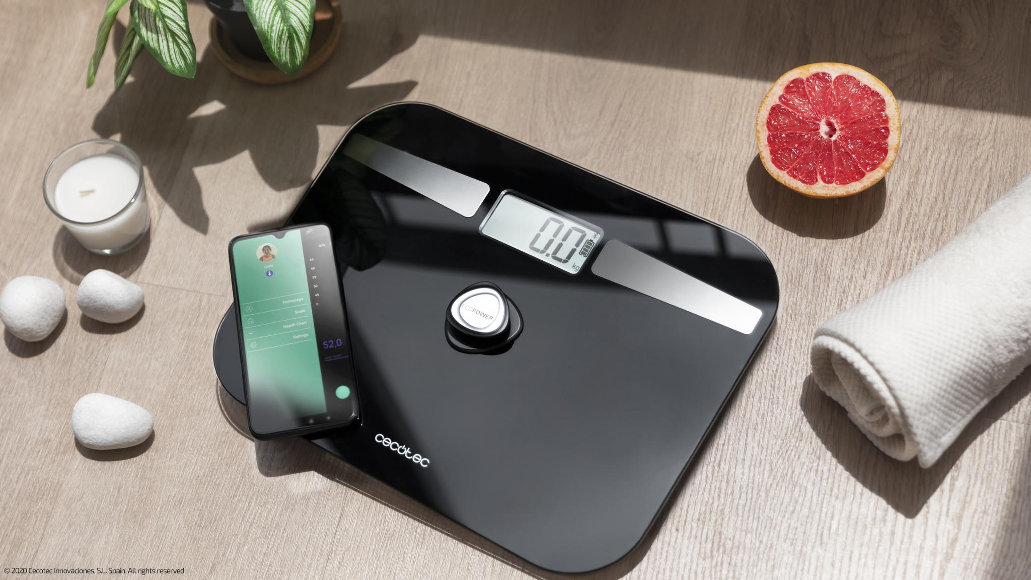 Balanza de baño digital negra con un teléfono inteligente, toalla, pomelo y vela en suelo de madera.