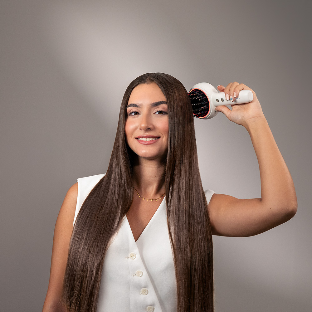 Una mujer sonriente con top blanco se cepilla el pelo castaño largo y liso con un cepillo blanco.