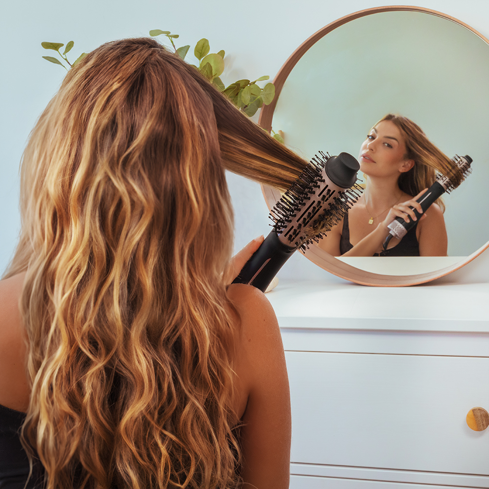 Una mujer se peina el cabello largo y castaño con un cepillo de aire caliente frente a un espejo.