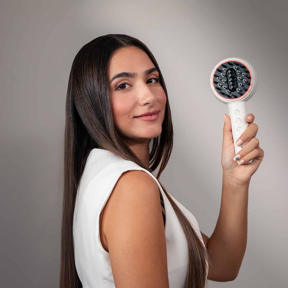 Una mujer con cabello castaño largo sonríe, sosteniendo un dispositivo para el cabello blanco sobre fondo gris.
