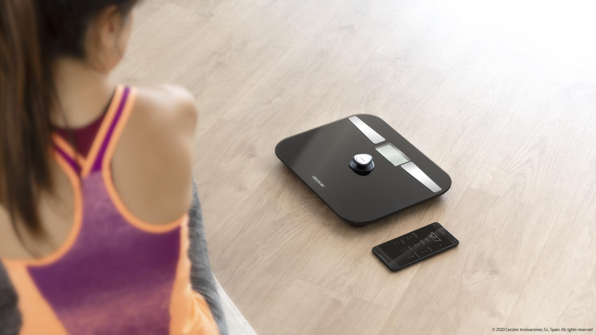 Una mujer en ropa deportiva se sienta en un suelo de madera junto a una báscula de baño digital y un teléfono inteligente.