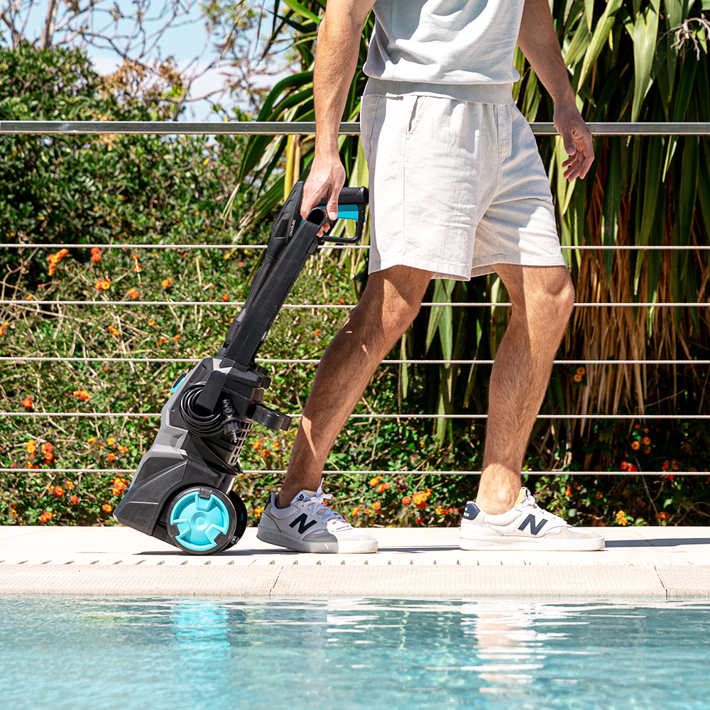 Un hombre con ropa clara de verano tira de una hidrolimpiadora negra y azul junto a una piscina.
