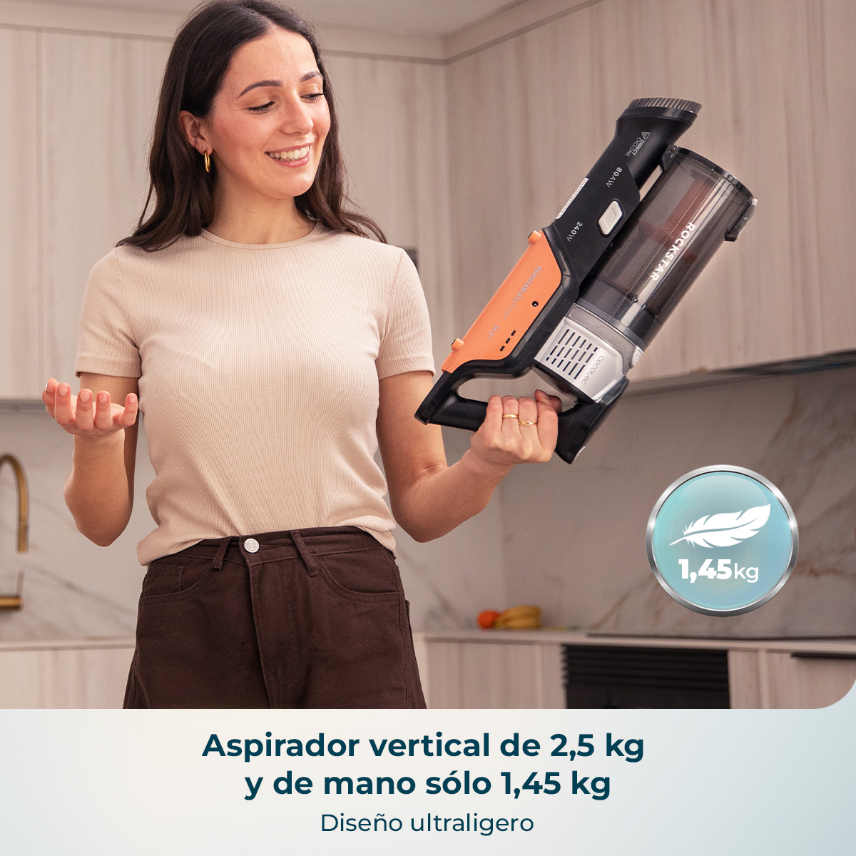 Una mujer sostiene una aspiradora de mano ligera negra y naranja en una cocina luminosa.