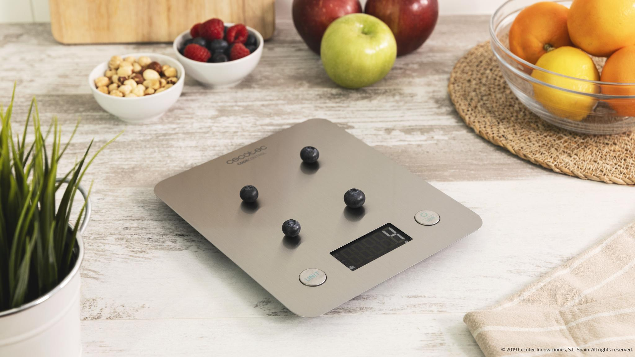 Una báscula de cocina de acero inoxidable sobre madera sostiene cuatro arándanos, rodeada de boles con frutos secos y fruta.