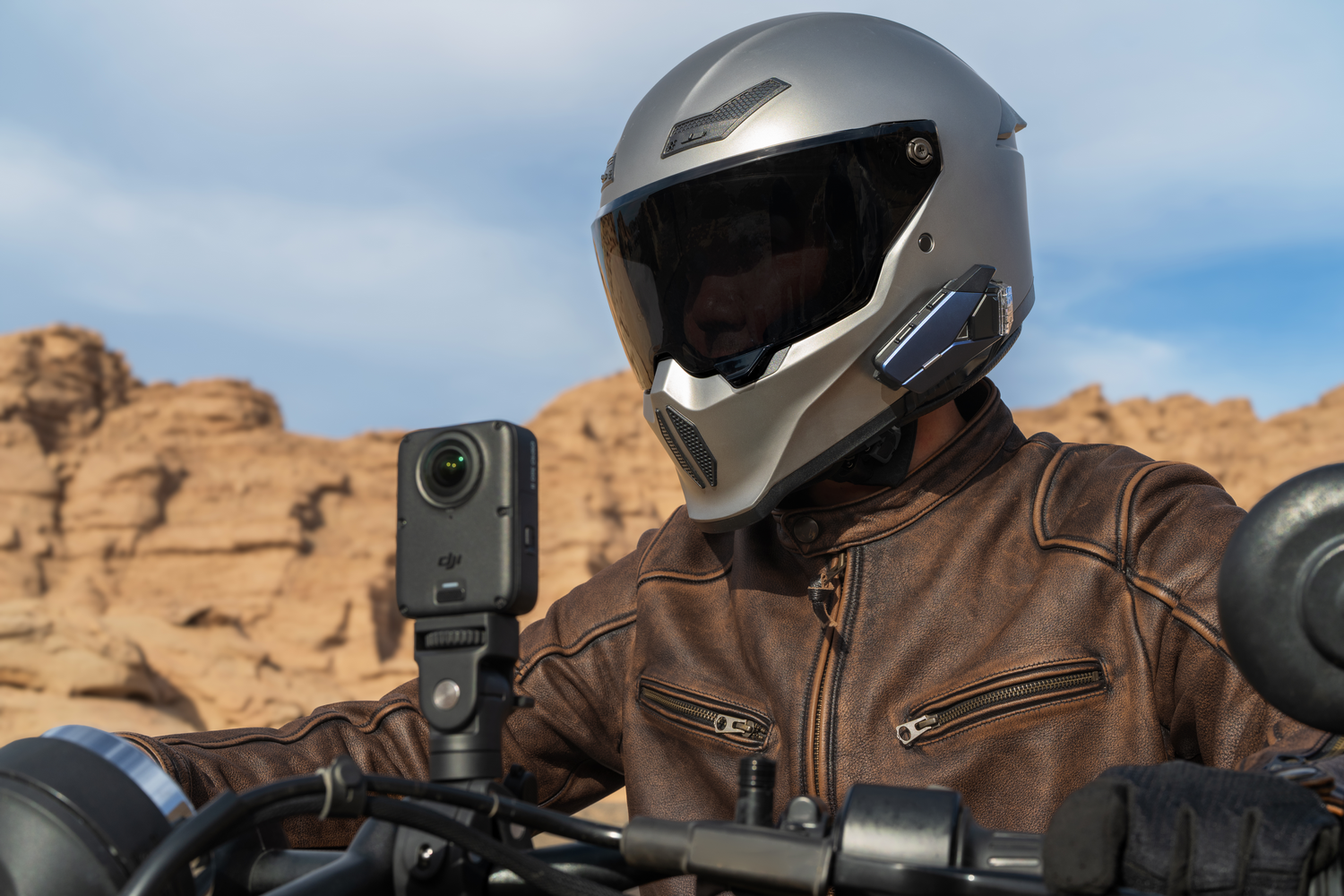 Motociclista con casco plateado y chaqueta de cuero marrón, con un paisaje montañoso al fondo.