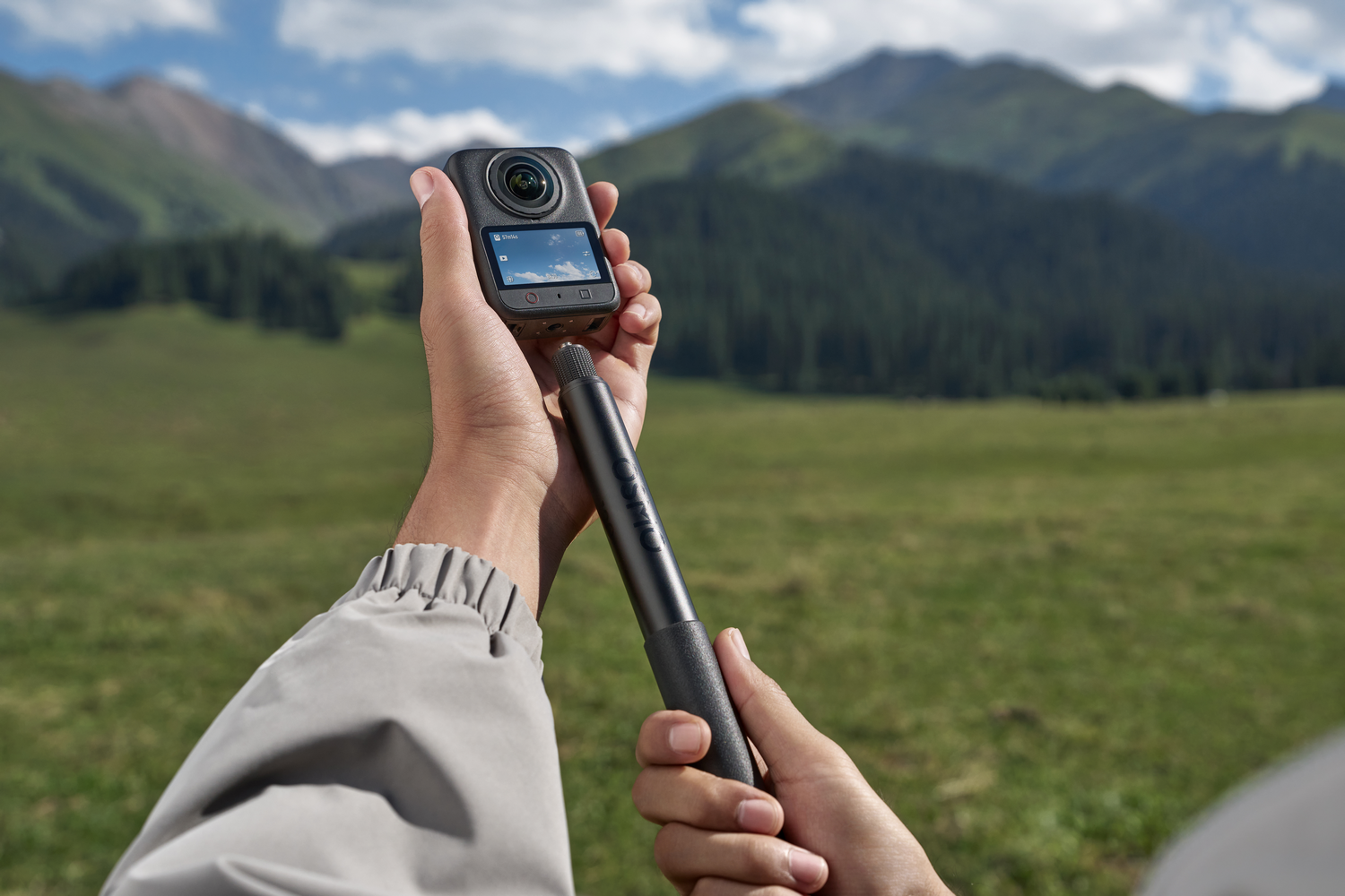 Una persona sostiene una cámara pequeña en un palo selfie frente a un paisaje montañoso.