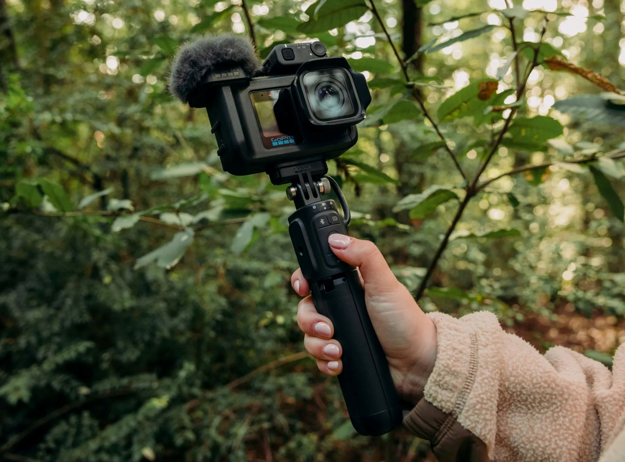 Eine Hand hält einen schwarzen GoPro-Handgriff mit Kamera und Mikrofon vor einem grünen Wald.